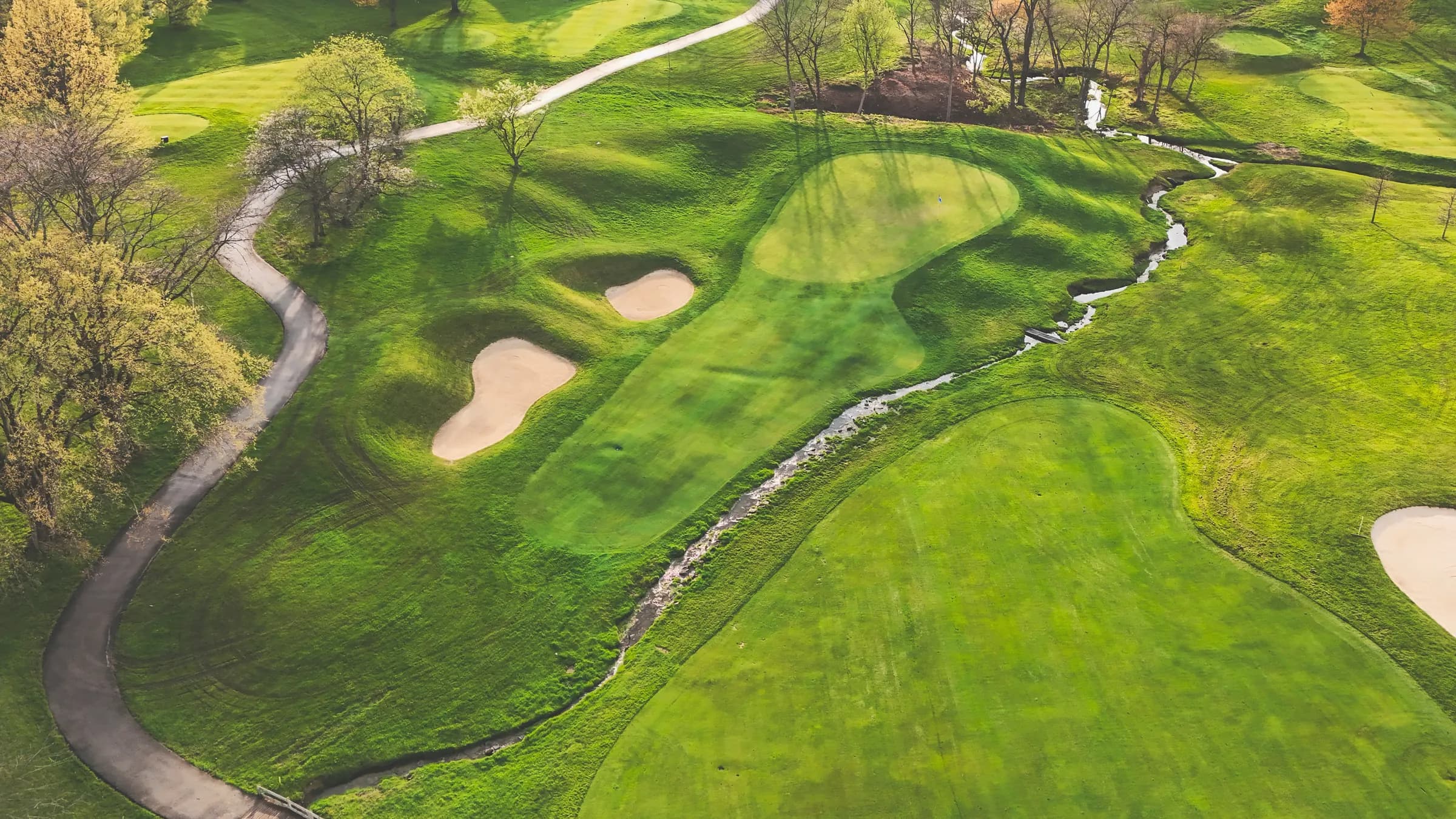 An aerial view of a golf hole showing the green, bunkers, cart path and a stream
